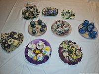 Nine miniature tea sets arranged on a white cloth, each with flower-themed patterns and displayed on various colored trays or plates.