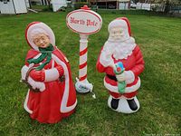Outdoor lawn display of three blow mold figures: Mrs. Claus, Mr. Claus, and a North Pole decorative sign. All situated on grassy lawn, showing condition and scale.