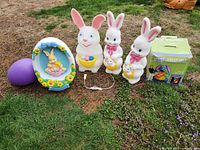 Photo showing five Easter-themed outdoor blow mold decorations arranged on grass, including three bunnies with baskets, one egg blow mold with bunny inside, and one large purple egg.