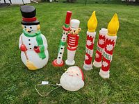 Outdoor Christmas blow molds arranged on grass showing snowman, Santa head, two red candles, bear soldier, and two clarinet player toy soldier figures