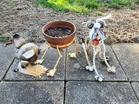 Two metal dog-shaped outdoor decorations on a paved surface, one sculpture and one planter with a rusty clay pot, showing signs of rust and outdoor weathering.