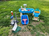 Blue toddler swing with yellow ropes, blue and green shopping cart, ball set with batters and tees, and blue water table with palm trees set on grass.