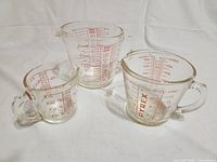 Three Pyrex glass measuring cups of different sizes arranged on a white background: 1 cup, 1 pint, and 1 quart sizes. Each has red measurement markings and the Pyrex name printed on the sides.