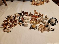 Photo showing a large assortment of small porcelain animal figurines arranged on a white surface. The animals are detailed with realistic and stylized features, including deer, skunks, lambs, an elephant, pandas, foxes, giraffes, ducks, a pig, a turtle, a unicorn, and small animal signs.