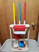 First photo view of Melissa & Doug wooden cleaning playset showing red, blue, and yellow handled tools on a wooden holder rack, mop head visible, mop handle shows cheek damage.