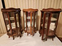 Three small wooden curio cabinets with glass panels, front doors open, placed on white surface against vertical blinds background.