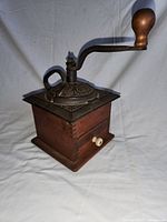 Side view of the vintage coffee grinder showing metal top, crank handle, wooden box body, and drawer with white knob.