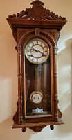 Front view of a vintage wooden wall clock with carved ornate top, side columns, glass door, Roman numeral clock face, and visible pendulum inside.