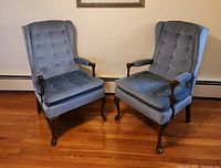 Pair of blue tufted Queen Anne style wing back chairs displayed side by side on a wooden floor against a plain wall, showing detailed upholstery and wooden legs and armrests.