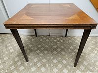 Front view of wooden folding card table showing square top with triangular wood inlay design and four tapered legs against a tiled floor.