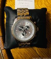 Front view of silver tone Akribos XXIV men's watch on black pillow. Shows round dial with three subdials, date window, and Arabic numerals.