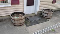 Two wooden barrel planters placed outside a house door on concrete. Each barrel filled with soil and natural debris, showing rusted metal bands and wood wear.