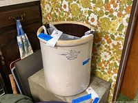 Front and side view of large stoneware crock with blue crown and number 10 mark, placed on wooden surface against floral wallpaper.