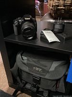 Photo showing Canon EOS digital camera with lens cap attached, battery charger, and battery on a shelf with a woven basket in background.