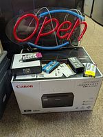 Neon 'Open' sign lit, multiple ink cartridges placed on top of Canon imageCLASS LBP122dw printer box