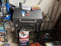 Full view of black Char Griller Traditional grill with side shelves, thermometer, bag of Kingsford charcoal in front, and grill utensils on side shelf.