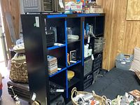 Two black cubical storage shelves standing side by side in a room with various items inside the compartments and around them.