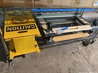 Two yellow caution signs on a metal shelf, one sign laying flat showing CAUTION TRIPPING HAZARD text and icon, two metal dollies with wheels below the shelf.