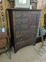 Front view of dark wooden dresser showing 5 drawers, one door, brass handles, and signs of wear