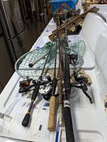 Fishing rods laid out with reels attached, green fishing net below, rod holder, and fishing vest in background.