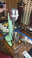 Full view of Hamilton Beach milk shake maker in green with chrome head, mixing cup, hand mixer, and Mouli grater on wood table.
