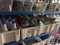 Metal shelving with four plastic bins containing disinfectant sprays, Lysol bottles, and cleaning supplies. Transparent bins on top shelf hold assorted light bulbs.