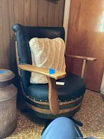 View of chair angled to display faux leather upholstery, partial pillow, wooden armrests, and oak wood barrel base with metal bands.