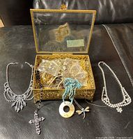 Open glass and brass tone jewelry box filled with assorted costume necklaces on a black surface. Three necklaces displayed outside the box including rhinestone and beaded styles.