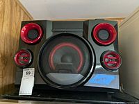Front view of the LG mini Hi-Fi speaker system showing one large central speaker with metal grille and red ring, and four smaller red-accented speakers around it.
