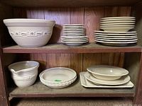 Wooden shelf with multiple stacks of off-white Longaberger plates, bowls, and serving dishes including small plates, dinner plates, small bowls, and large serving bowls