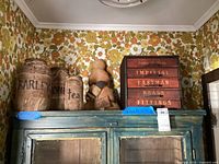 Wide view of vintage sealed jars, plush bee toy, and metal fittings box atop a cabinet with floral wallpaper background.