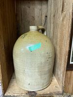 Front and side views of a vintage stoneware jug approximately 15 inches tall with a single handle and narrow spout, light brown glaze with speckled texture, showing natural wear and markings.