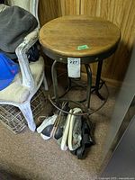 Side view of vintage industrial stool showing wooden top, metal legs with footrest ring, surrounded by clothes on floor and adjacent chair.
