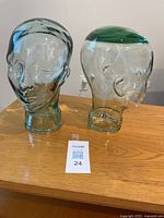 Two clear glass head sculptures placed on wooden table with maxsold card in front. Shows front view of each head and their height scale.