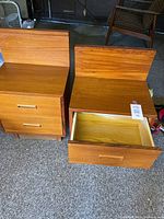 Pair of teak wood nightstands with two drawers each, one drawer open revealing interior, showing wood grain and horizontal handles.