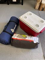 Photo shows blue rolled Woods sleeping mat, red and white Coleman cooler, and two packed camping gear bags including the tent and sleeping bag.