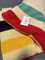 Portion of folded blanket showing red, yellow, green and black stripes on cream wool with maxsold tag.