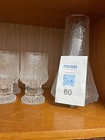 Tall clear textured glass vase with characteristic cracked ice pattern next to stemmed glasses on wooden shelf.
