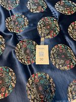 Close-up of navy blue brocade fabric showing circular oriental motifs in gold, red, turquoise, and black thread on smooth shiny surface with MaxSold tag