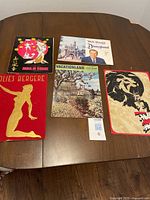 Photo showing all five pieces laid out on table: Moulin Rouge poster, Disneyland booklet with Walt Disney image, Vacationland booklet, Folies Bergère poster, and another vintage poster.