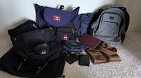 Full view of all bags arranged on floor showing variety including Matein grey backpack, navy and black bags with Canada flag emblems, and vintage brown leather Roots crossbody bag.