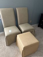 Two beige linen fabric slipper chairs with skirted bottoms and one matching yellow fabric ottoman on carpeted floor against blue wall.