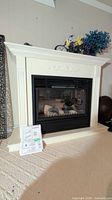 White electric fireplace with decorative molding and black front grill, floral design on upper front panel, placed on carpeted floor with instruction manual leaning against base.