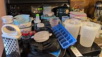 Overview of glass bakeware including Corning casseroles with Amish Butterprint pattern, clear and white plastic food storage containers of various sizes with matching lids, white pitchers, metal grater, blue ice cube tray, measuring cups, and black insulated lunch bag.
