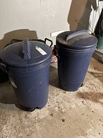 Two navy Rubbermaid Roughneck garbage cans with lids and wheels in a garage setting. Left can shows a sticker on the lid. Both cans exhibit dirt and signs of use.