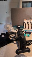 Lot arranged on carpet including foot spa device, small white desk fan, three boxed Moen handgrips, small blue weights, and black back brace strap in foreground.