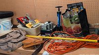Overview of workbench surface with leather tool belt with metal rings, orange extension cord, and various containers with tools and accessories including paint tools and a hand pump.