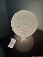 Lamp turned on, showing light diffusion through woven spherical shade and light pattern on surface below.