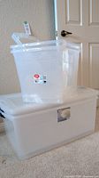 Two clear plastic Sterlite totes stacked, the bottom tote is a larger size with a lid, and the top tote is a smaller Sterlite tote with a lid.