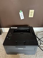 Front view of black Brother printer powered off next to a wall outlet.